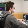 Individual seated at a computer desk, focused on the screen with a keyboard and monitor in view.